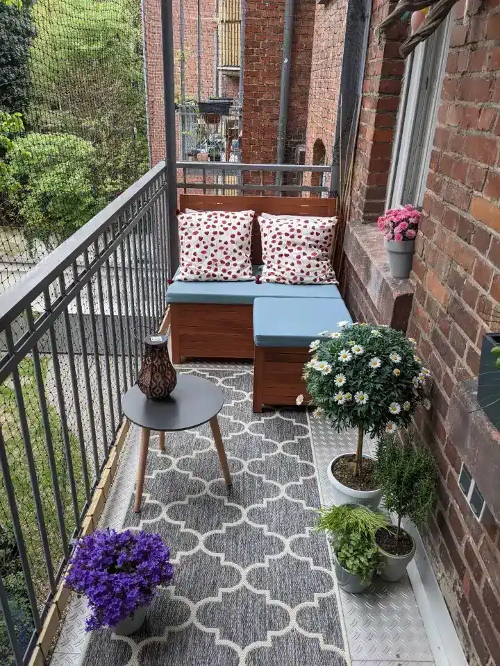 Klein balkon inrichten: 8 slimme en stijlvolle ideeën Overzichtelijk balkon met bloempotten op een smalle plantenstandaard en een ruimtebesparende balkonbank met hocker voor groen en functionaliteit